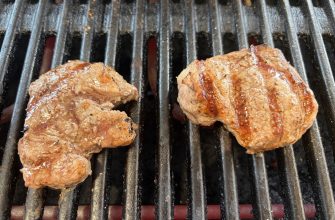Grilled beef pieces on a barbecue grill with dark grill bars and sizzling juices visible on the meat.